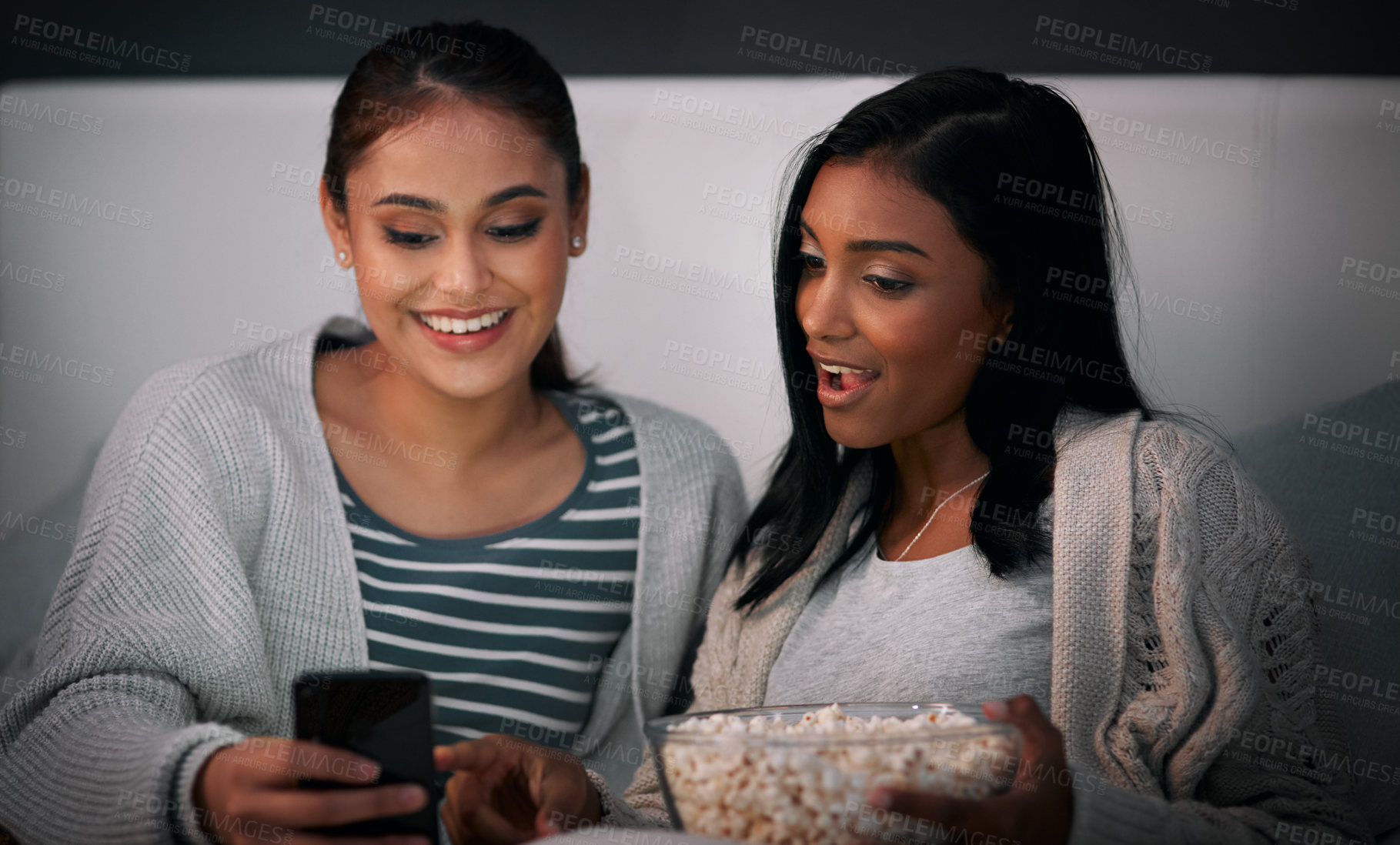 Buy stock photo Shot of a young woman showing her friend something on her cellphone while sitting together on a bed