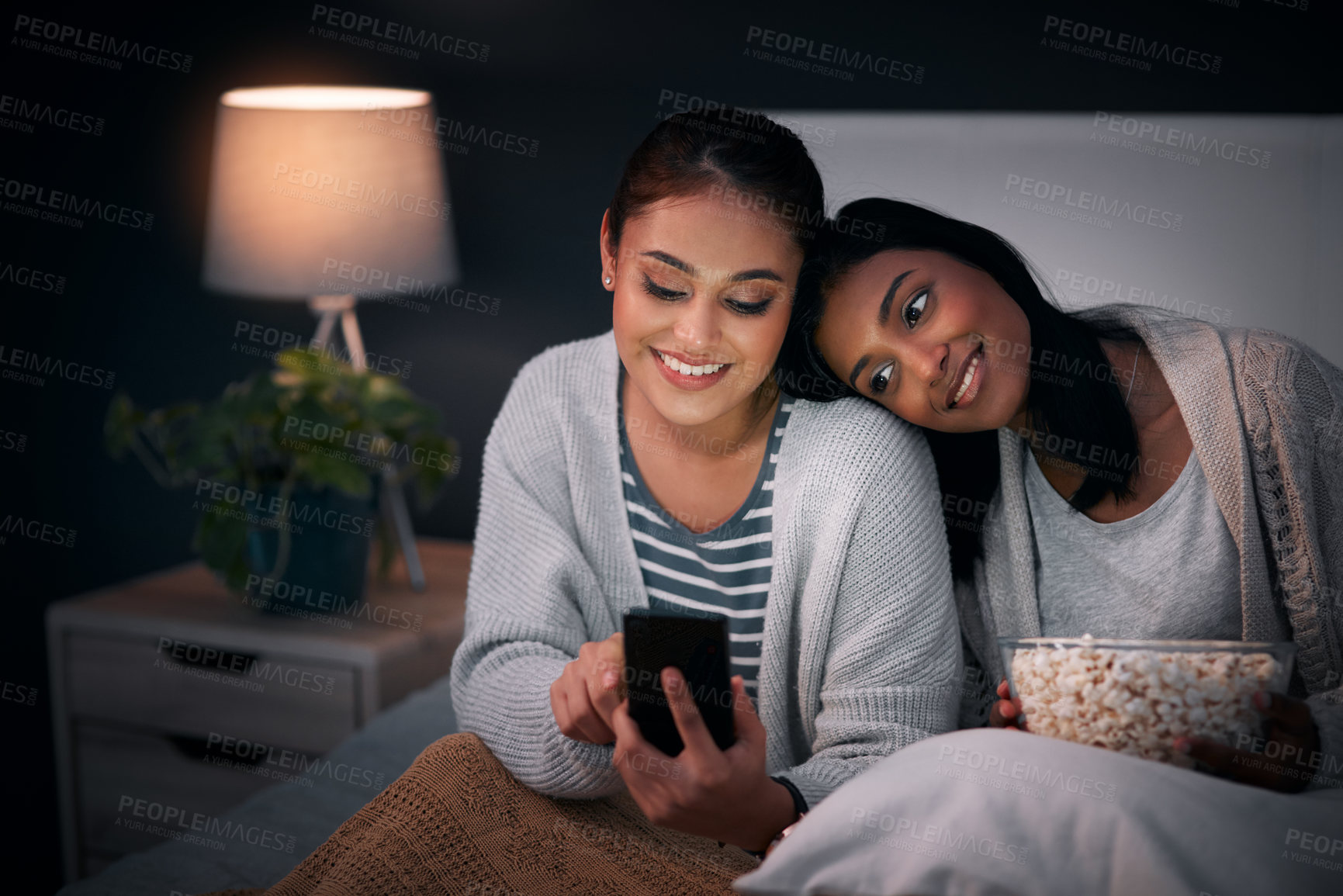 Buy stock photo Shot of a young woman showing her friend something on her cellphone while sitting together on a bed