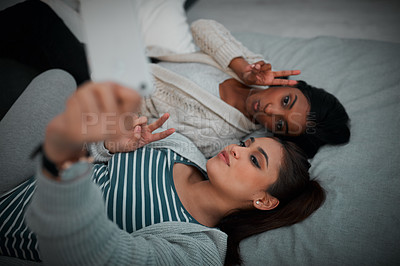 Buy stock photo Shot of two young women lying taking selfies while lying on a bed