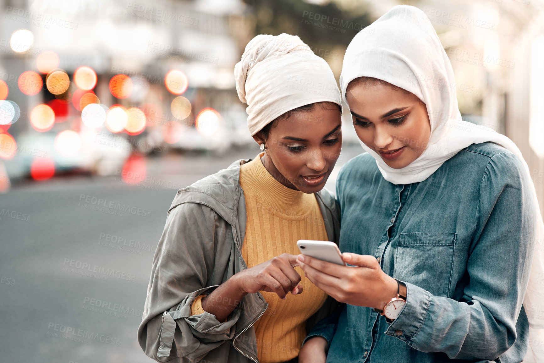 Buy stock photo Cropped shot of two attractive young women wearing headscarves and standing together while using a cellphone in the city