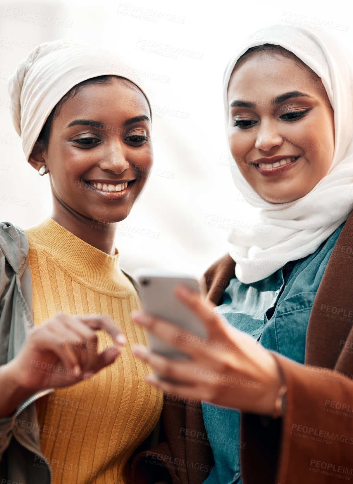 Buy stock photo Cropped shot of two attractive young women wearing headscarves and standing together while using a cellphone in the city