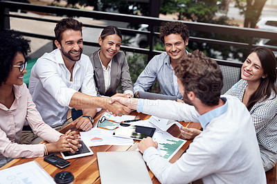 Buy stock photo Cropped shot of two handsome young businessmen shaking hands while sitting with their colleagues outdoors
