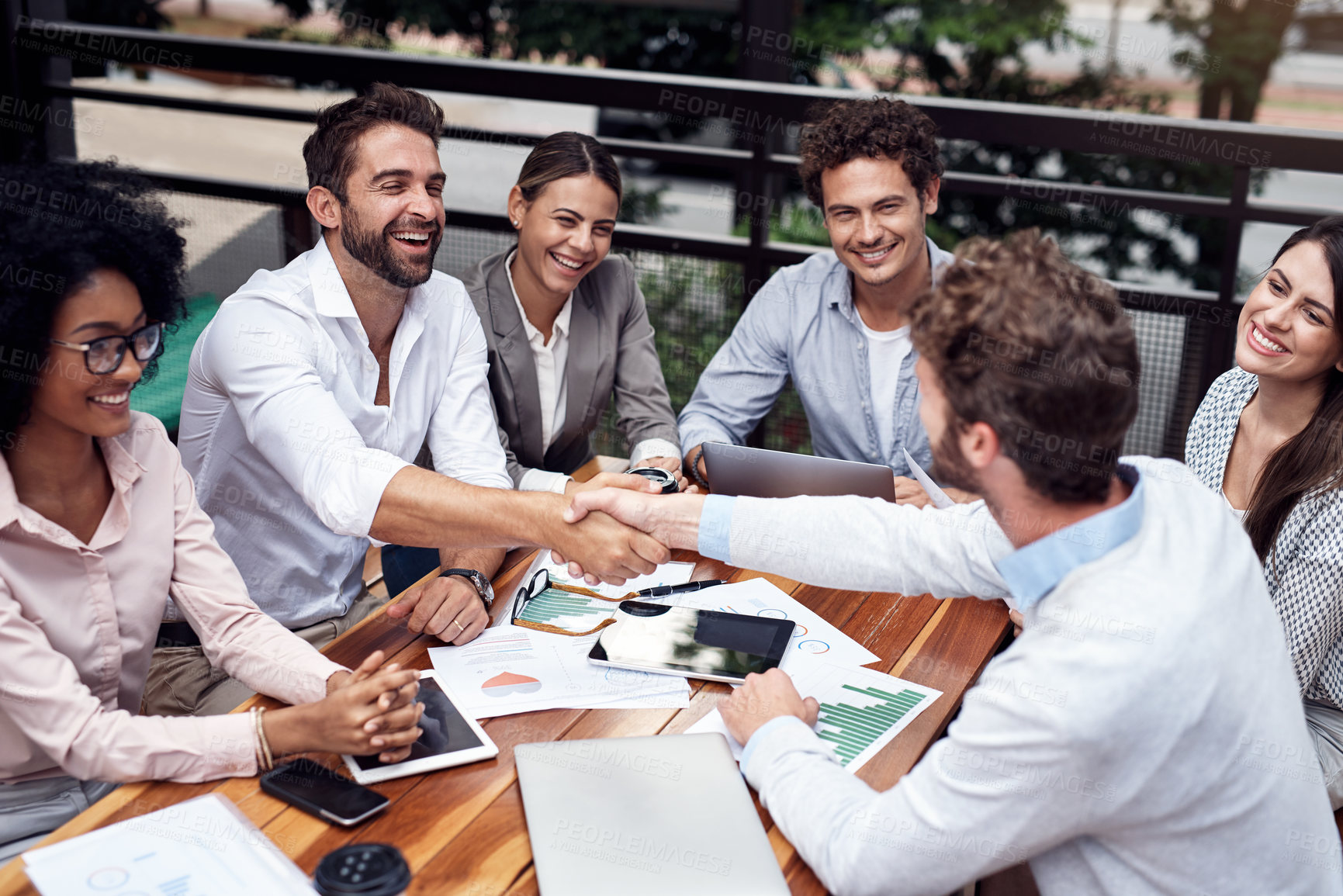 Buy stock photo Cropped shot of two handsome young businessmen shaking hands while sitting with their colleagues outdoors