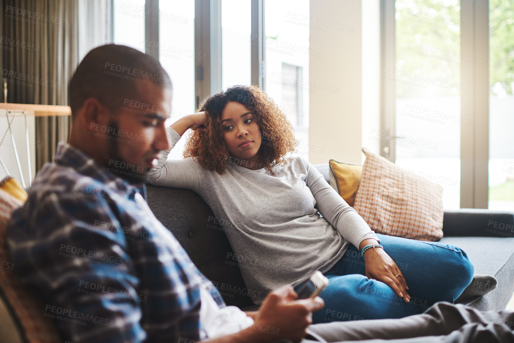 Buy stock photo Cropped shot of an attractive young woman sitting on the sofa and watching her husband use his cellphone