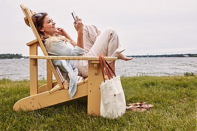 Buy stock photo Shot of an attractive young woman using a smartphone while relaxing next to the lake