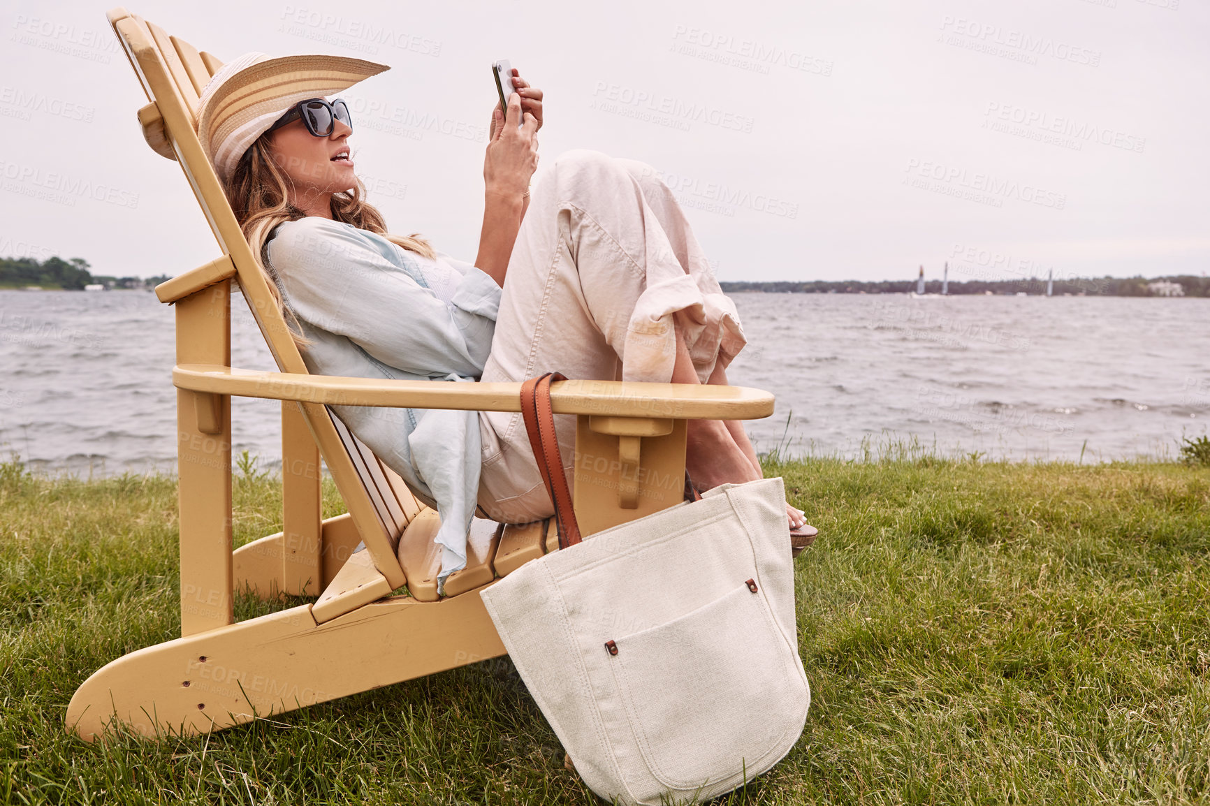 Buy stock photo Shot of an attractive young woman using a smartphone while relaxing next to the lake