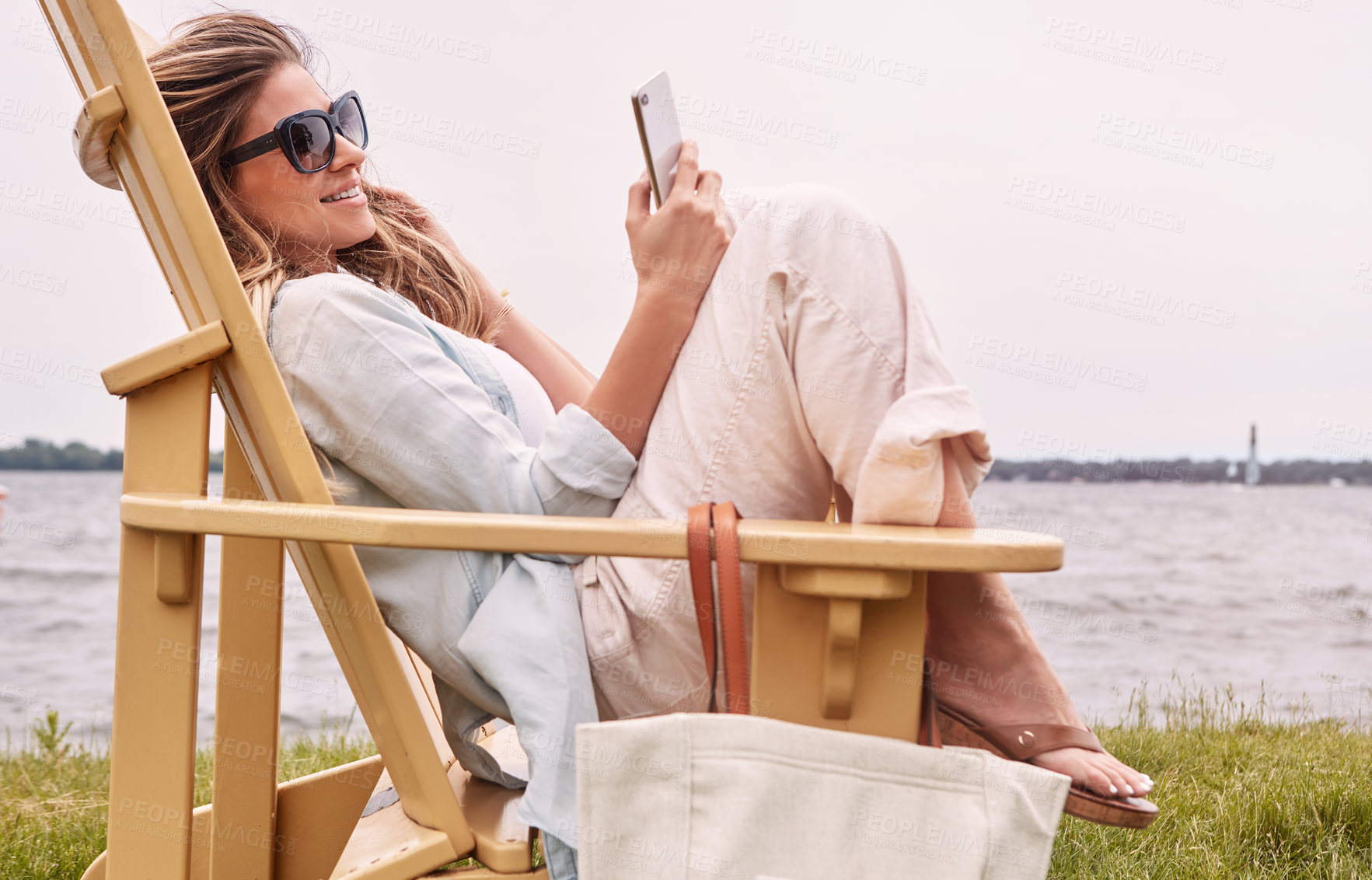 Buy stock photo Shot of an attractive young woman using a smartphone while relaxing next to the lake