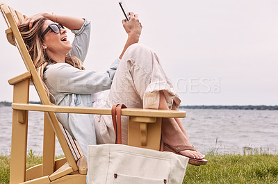 Buy stock photo Shot of an attractive young woman using a smartphone while relaxing next to the lake