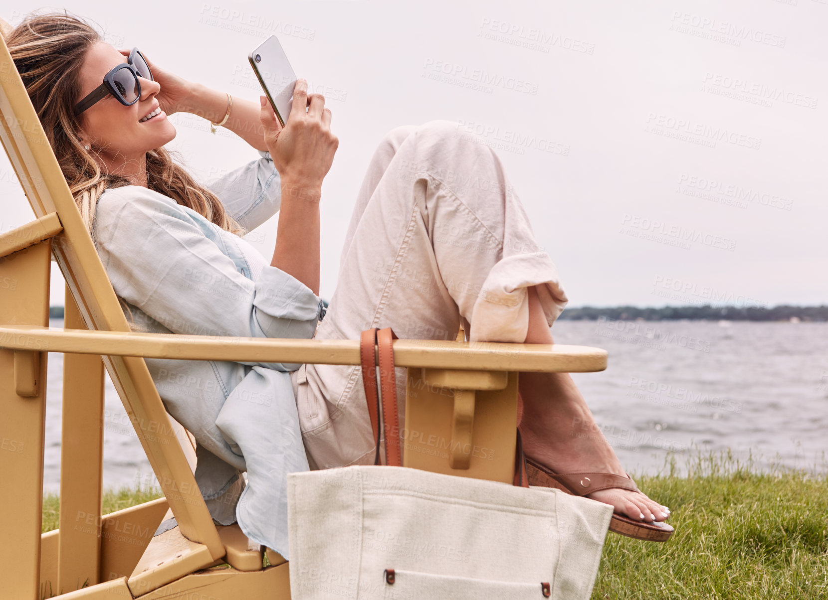 Buy stock photo Shot of an attractive young woman using a smartphone while relaxing next to the lake