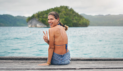 Buy stock photo Cropped portrait of an attractive young woman sitting on the edge of a boardwalk and making a peace sign gesture