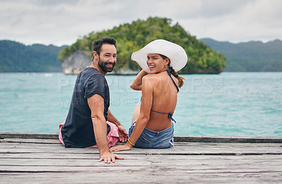 Buy stock photo Cropped portrait of a happy young couple sitting together on the boardwalk overlooking the ocean during vacation