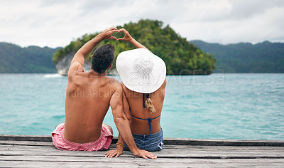 Buy stock photo Heart hands, back and couple by a ocean while on summer, paradise and tropical vacation. Romance, dock and young man and woman relaxing on wood pier with love shape gesture on holiday or weekend trip
