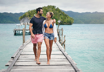 Buy stock photo Full length shot of a happy young couple walking arm in arm down a boardwalk during a vacation together