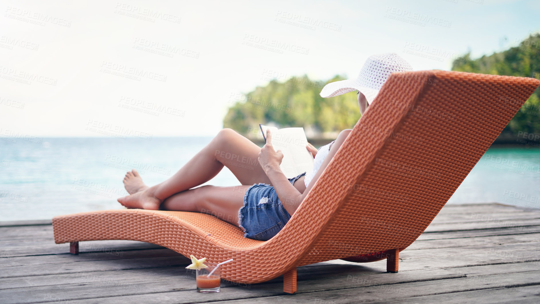 Buy stock photo Full length shot of an attractive young woman sitting on a sun lounger and reading a book during a vacation