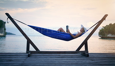 Buy stock photo Full length shot of an attractive young woman sitting alone in a hammock and reading a book during a vacation