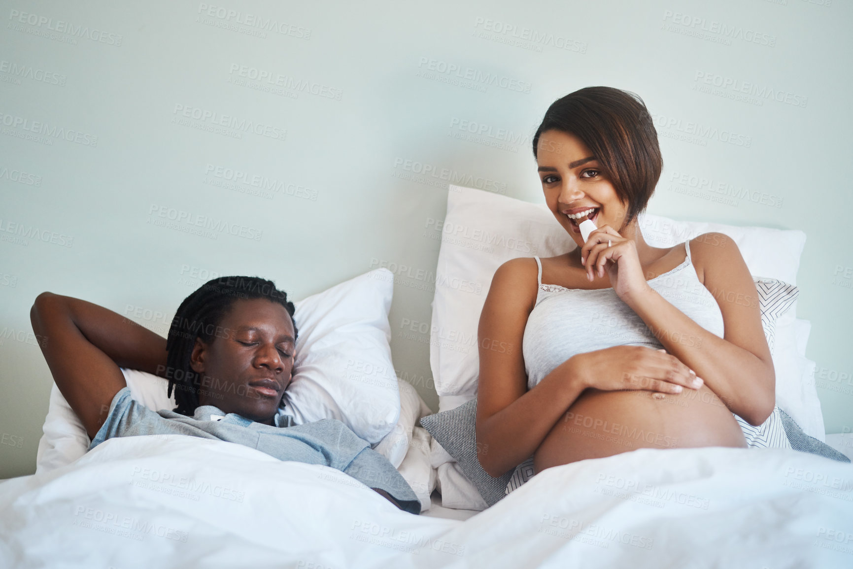 Buy stock photo Shot of an attractive young pregnant woman enjoying a chocolate bar while lying in bed with her husband