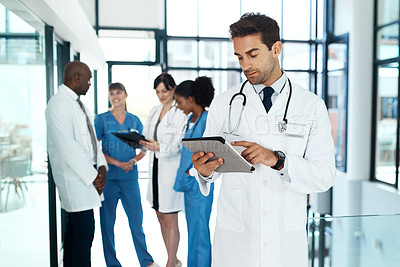 Buy stock photo Shot of a young doctor using a digital tablet in a hospital with his colleagues in the background