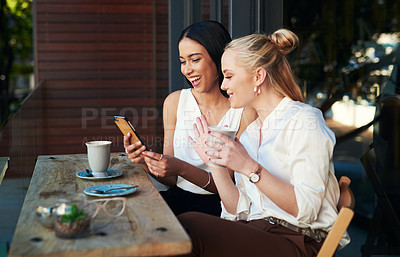 Buy stock photo Shot of two young women looking at something on a cellphone while sitting at a cafe