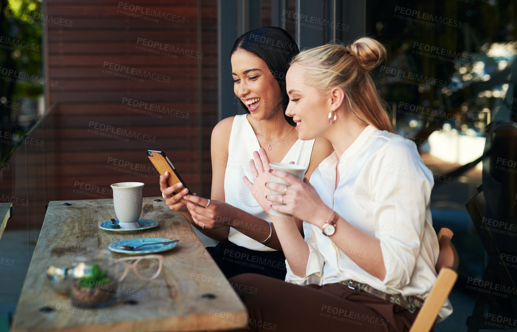 Buy stock photo Shot of two young women looking at something on a cellphone while sitting at a cafe