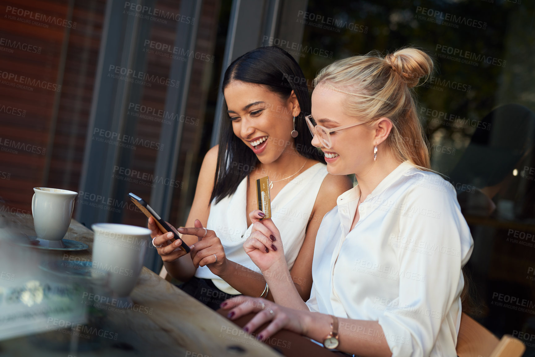 Buy stock photo Shot of a woman using a cellphone while her friend holds up a credit card while sitting together