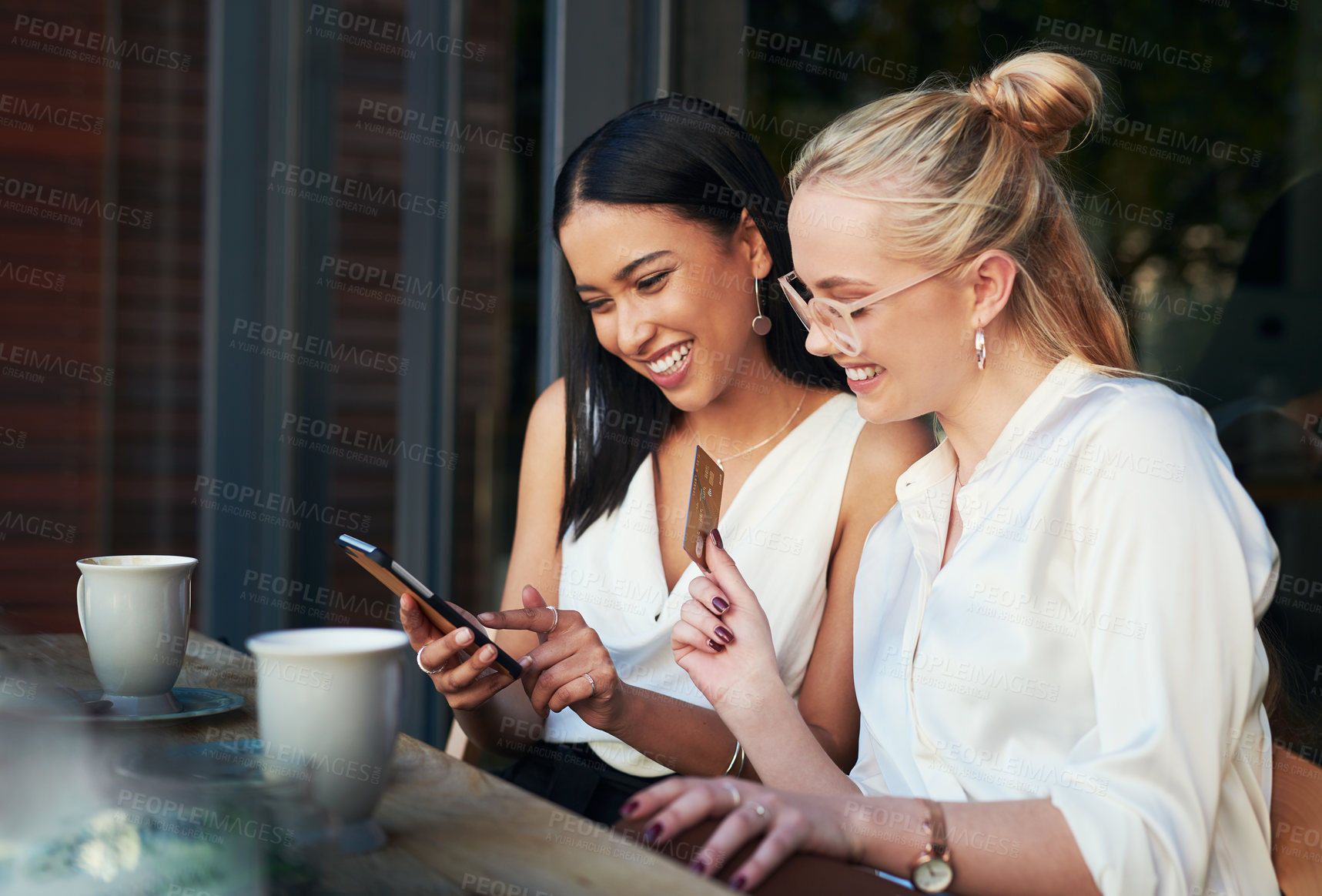 Buy stock photo Shot of a woman using a cellphone while her friend holds up a credit card while sitting together