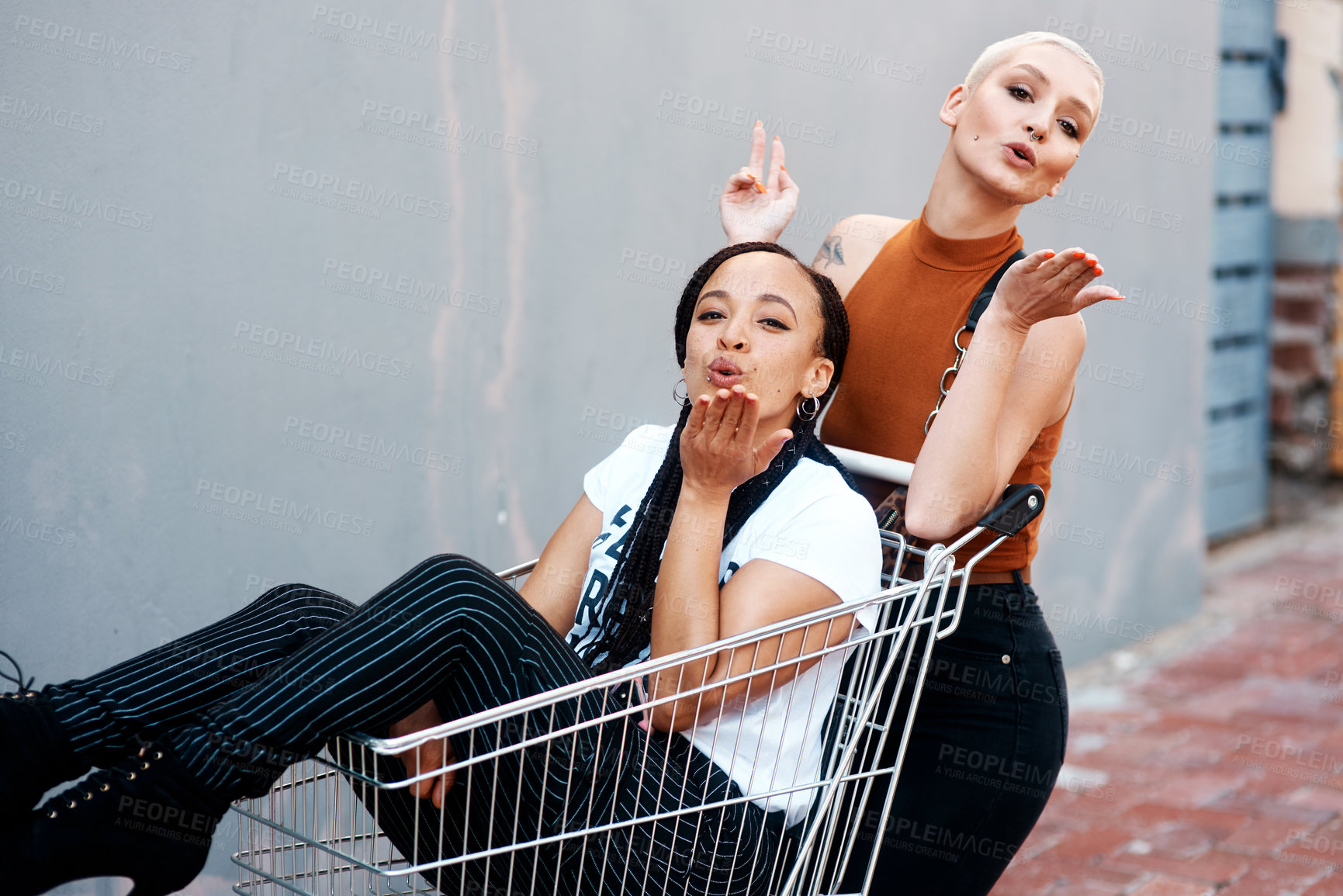 Buy stock photo Cropped portrait of two cheerful young girlfriends blowing kisses while playing around with a shopping cart outdoors