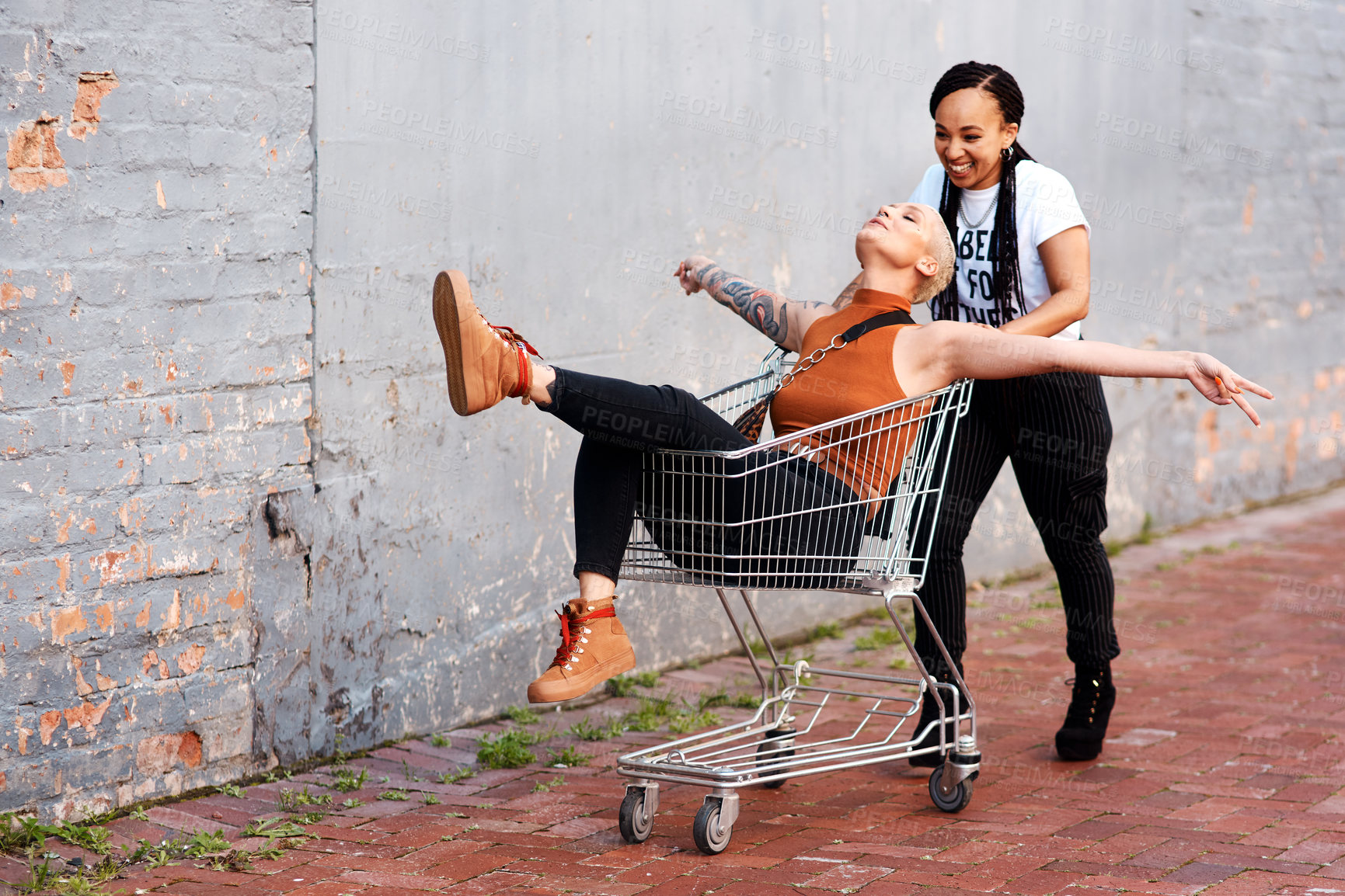 Buy stock photo Full length shot of an energetic young woman pushing her female friend in a shopping cart outdoors
