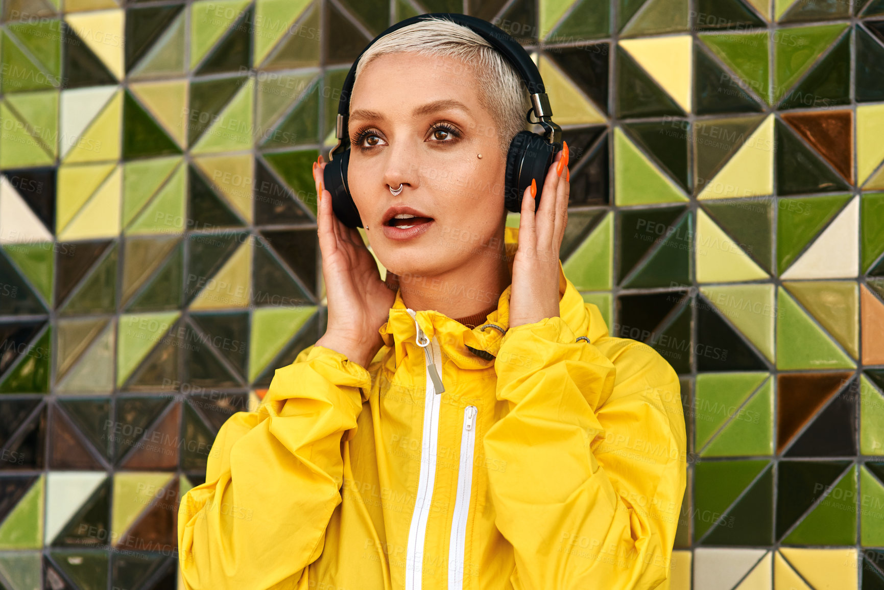 Buy stock photo Cropped shot of an attractive young woman standing against a tiled wall and listening to music through headphones