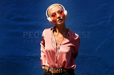 Buy stock photo Cropped portrait of an attractive young woman standing against a blue wall and listening to music through headphones
