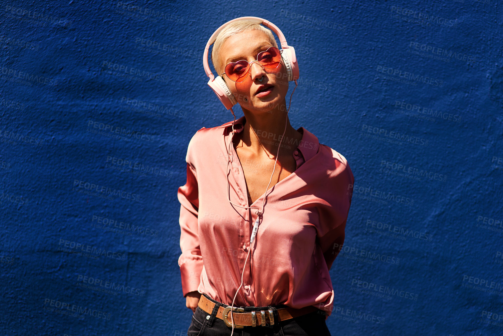 Buy stock photo Cropped portrait of an attractive young woman standing against a blue wall and listening to music through headphones