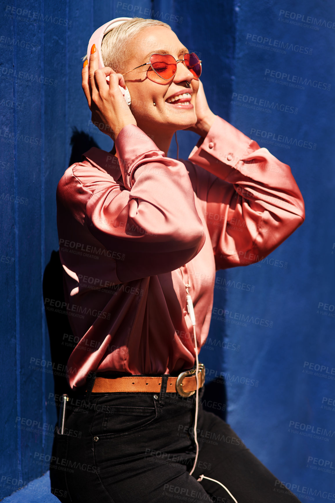 Buy stock photo Cropped shot of an attractive young woman standing against a blue wall and listening to music through headphones