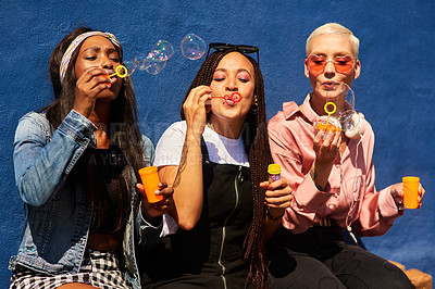 Buy stock photo Cropped shot of three attractive young women sitting against a blue wall together and bonding by blowing bubbles