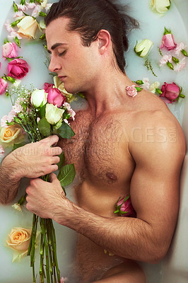 Buy stock photo High angle shot of a handsome young man holding a bouquet while lying in a bathtub full of milky water and flower petals at home
