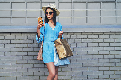 Buy stock photo Cropped shot of an attractive young woman using a smartphone while holding shopping bags against a brick wall