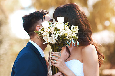 Buy stock photo Shot of a bride and groom covering their faces with a bouquet while kissing