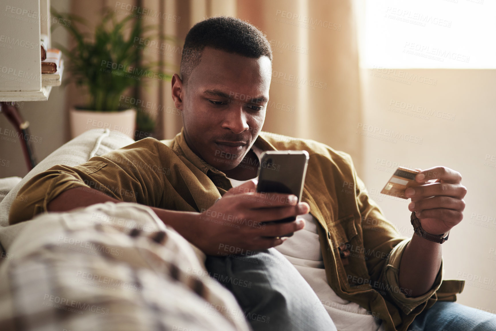 Buy stock photo Cropped shot of a handsome young man using a smartphone and a credit card to shop online while sitting on his couch at home