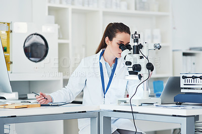 Buy stock photo Cropped shot of a focused young female scientist looking through a microscope and making notes inside of a laboratory