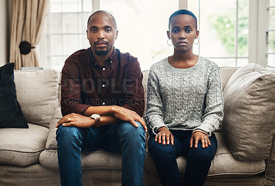 Buy stock photo Portrait of a young couple looking serious and sitting alongside each other on a sofa at home
