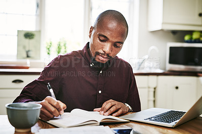Buy stock photo Shot of a handsome man going over paperwork and using his laptop while working from home during the day