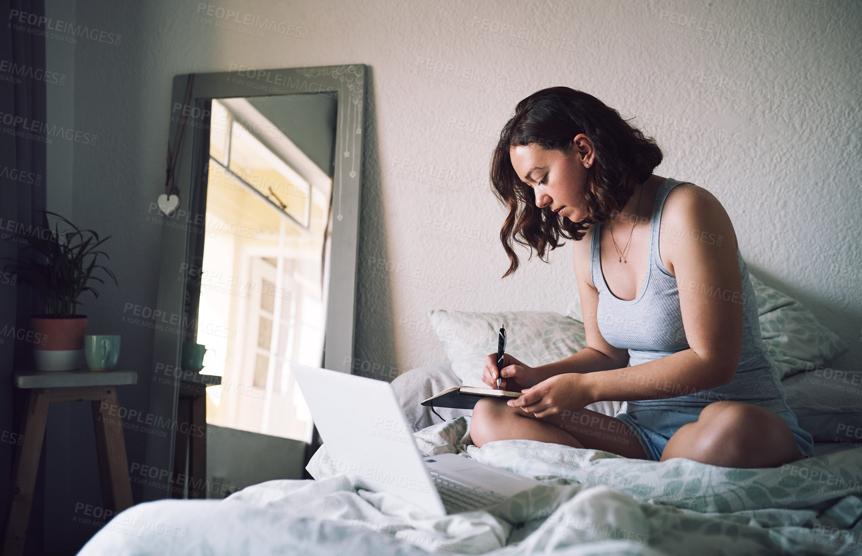 Buy stock photo Full length shot of an attractive young woman writing in a notebook while using a laptop in her bedroom at home