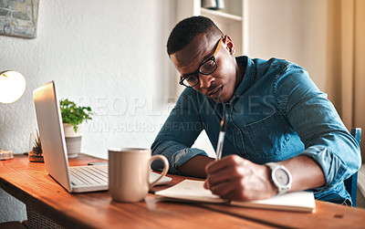 Buy stock photo African american entrepreneur writing in a notebook and planning a project strategy. Young man remote working from home with a laptop at his startup business while scheduling appointments in a book