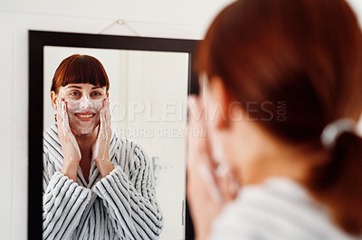 Buy stock photo Cropped shot of an attractive young woman giving herself a facial treatment while standing in front of the mirror in her bathroom at home