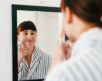 Buy stock photo Cropped shot of an attractive young woman applying moisturizer on her face while standing in front of the mirror at home