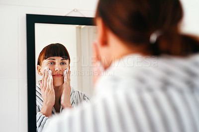 Buy stock photo Cropped shot of an attractive young woman applying beauty treatment on her face while standing in front of the mirror at home