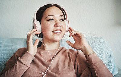 Buy stock photo Cropped shot of an attractive young woman sitting on her home sofa alone and listening to music through headphones