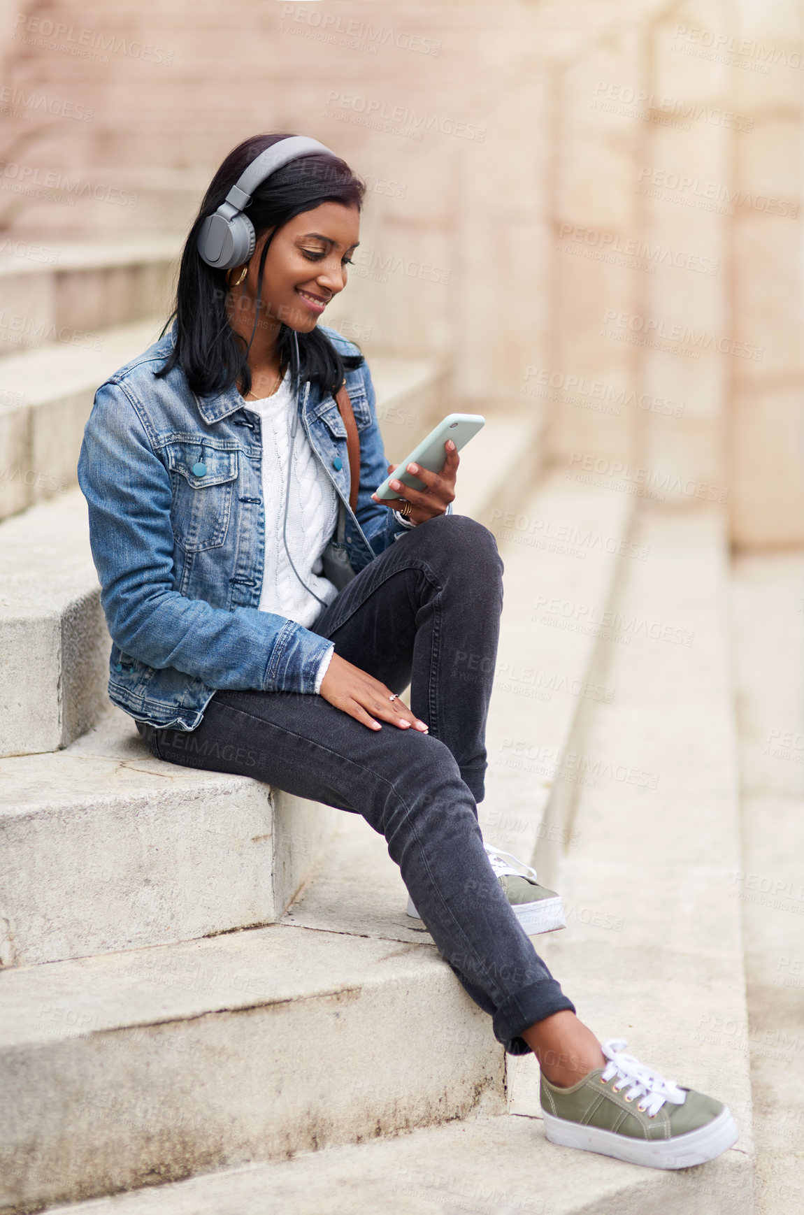 Buy stock photo Shot of a young woman wearing headphones while using her cellphone out in the city