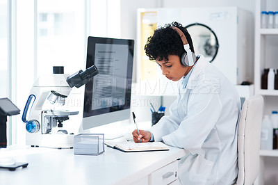 Buy stock photo Shot of a young scientist wearing headphones while writing notes and working on a computer in a lab