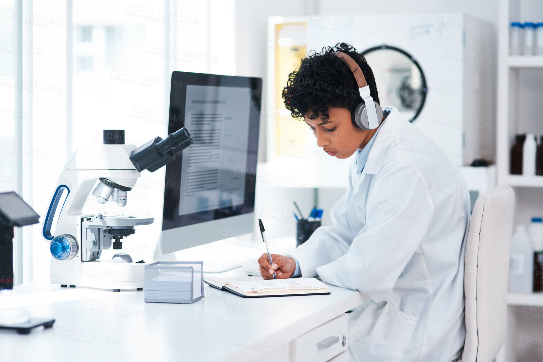 Buy stock photo Shot of a young scientist wearing headphones while writing notes and working on a computer in a lab