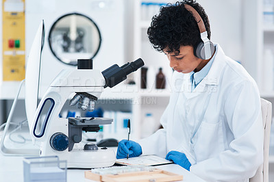 Buy stock photo Shot of a young scientist wearing headphones while writing notes and using a microscope in a lab