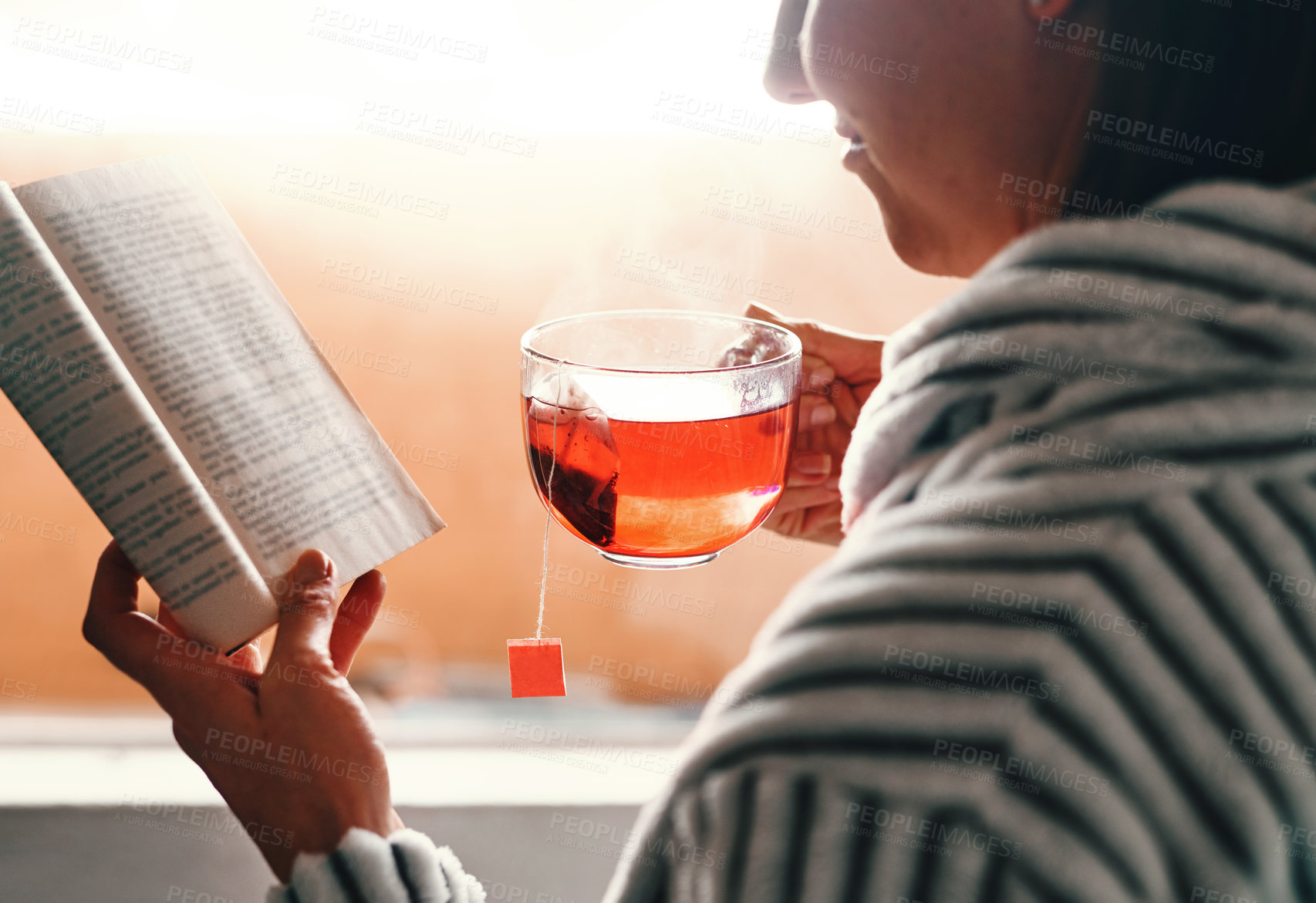 Buy stock photo Cropped shot of an unrecognizable young woman reading a book while drinking a cup of tea at home